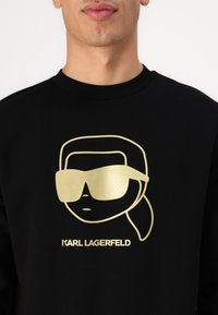 Schwarzer Sweatshirt mit einem goldenen Grafik eines Charakters mit Sonnenbrille, neben dem Text "KARL LAGERFELD" in fetter, moderner Schrift.