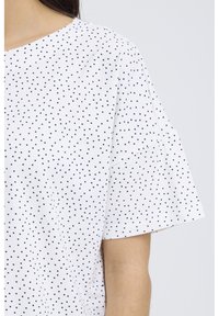 Biela krátka košeľa s malými čiernymi bodkami, zobrazená od ramena po stred paže na osobe s viditeľnými tmavými vlasmi.