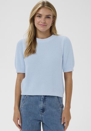 Tânără cu păr lung blond, purtând cercei aurii în formă de cerc, un pulover tricotat cu mâneci scurte, de culoare albastru deschis, și blugi bleu cu talie înaltă.