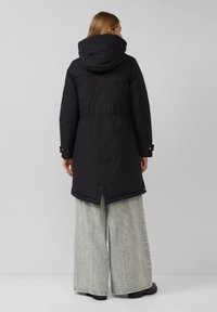 Schwarze Parkajacke mit Kapuze, elastischem Bund und einem längeren Rückensaum. Wird über grauen weit geschnittenen Hosen mit schlichtem Design getragen.