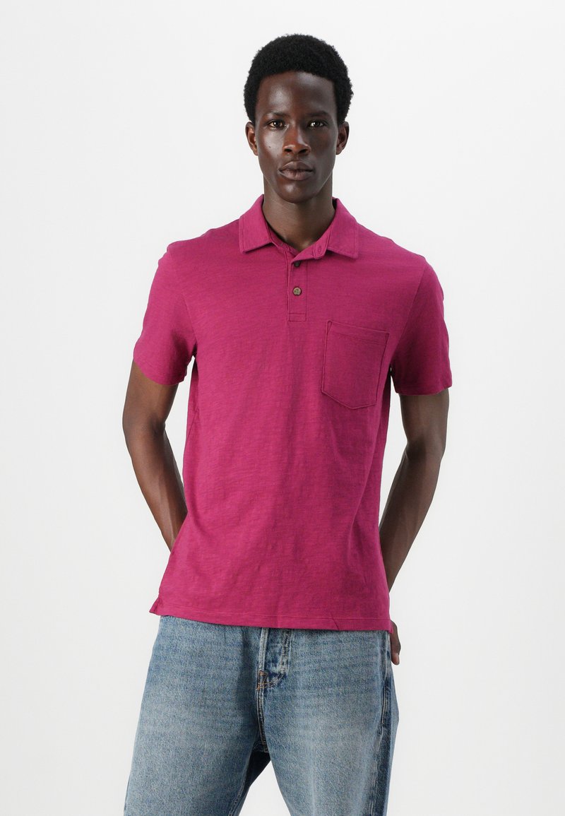 T-shirt polo en magenta vif avec des manches courtes, un plastron à deux boutons et une poche sur la poitrine. Fabriqué en tissu texturé léger.