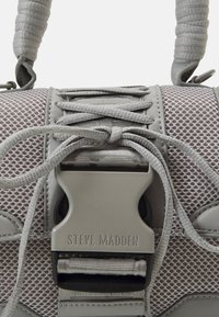 Sac à main gris avec un design en maille, doté d'une boucle proéminente avec le logo "STEVE MADDEN" et de poignées texturées pour une prise en main et un style.