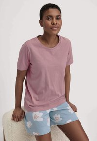 Kurzärmliges, pinkes Baumwoll-T-Shirt, kombiniert mit hellblauen Shorts mit weißen Blumenmuster. Das Model sitzt auf einer strukturierten Oberfläche.
