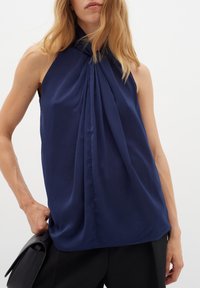 Marinblå ärmlös blus med hög halsringning och symmetriska veck fram. Mjuk tyg, avslappnad passform och subtil lyster.