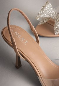 Sandale beige à talons hauts et bout ouvert, avec une bride arrière en daim et une bande transparente, ornée d'un nœud en strass brillant sur l'autre chaussure.