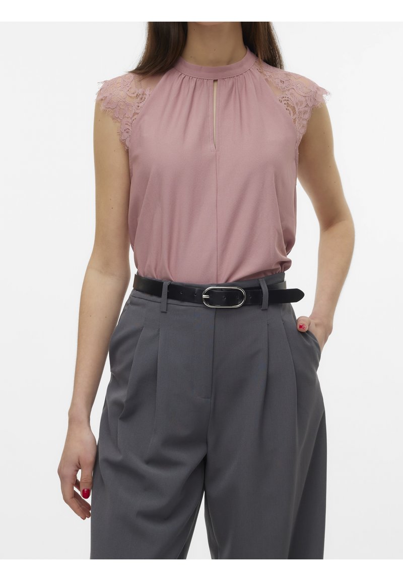 Femme portant un top en dentelle rose sans manches, rentré dans un pantalon gris taille haute avec une ceinture noire, debout avec une main dans la poche.