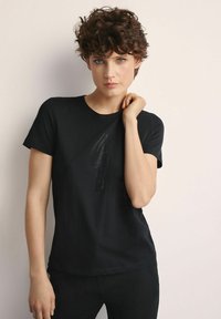 Kobieta z krótkimi kręconymi włosami, ubrana w czarną koszulkę z delikatnym teksturowanym wzorem, pozująca z jedną ręką przy szyi na jednolitym tle.