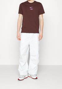 Brązowy bawełniany t-shirt z purpurowym logo Nike i tekstem, w połączeniu z białymi spodniami cargo i czarno-czerwonymi trampkami, stojący na neutralnym tle.