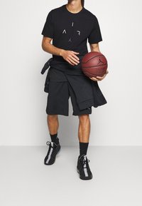 Černé tričko Jordan s logem "AIR", v kombinaci s černými kraťasy, držící basketbal. Černé sportovní boty s šedými akcenty.