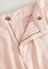 Pantalon rose clair avec fermeture éclair ouverte et deux boutons en écaille visibles à la taille sur fond uni.