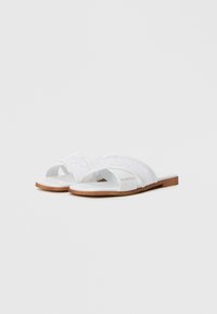 Hvite sandaler med kryssede snørestropper, en glatt såle og treinnlegg. Har branding på siden for identifikasjon.