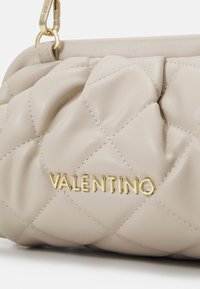 Beige kožená kabelka s prošívaným vzorem, se zlatými kovovými doplňky a embosovaným logem "VALENTINO" na přední straně. Měkká textura a strukturovaný tvar.