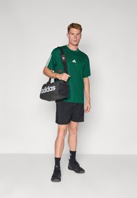 Zwarte sporttas met wit "adidas" logo, vastgehouden door een persoon in een groen mesh shirt, zwarte shorts en zwarte sneakers, staande op een effen achtergrond.