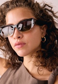 Cat-Eye-Sonnenbrille mit transparentem schwarz-braunem Gestell, dunklen Gläsern und glänzendem Finish. Wird mit einem einfachen, neutralfarbenen Outfit getragen.