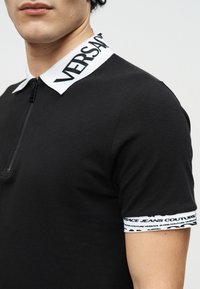 Άνδρας που φοράει μαύρο πόλο πουκάμισο με λευκό γιακά και μανσέτες, με σήμανση Versace Jeans Couture.