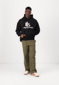 Felpa nera con logo bianco, pantaloni cargo olive e scarpe bianche con righe rosse. Posizione eretta su sfondo bianco uniforme.