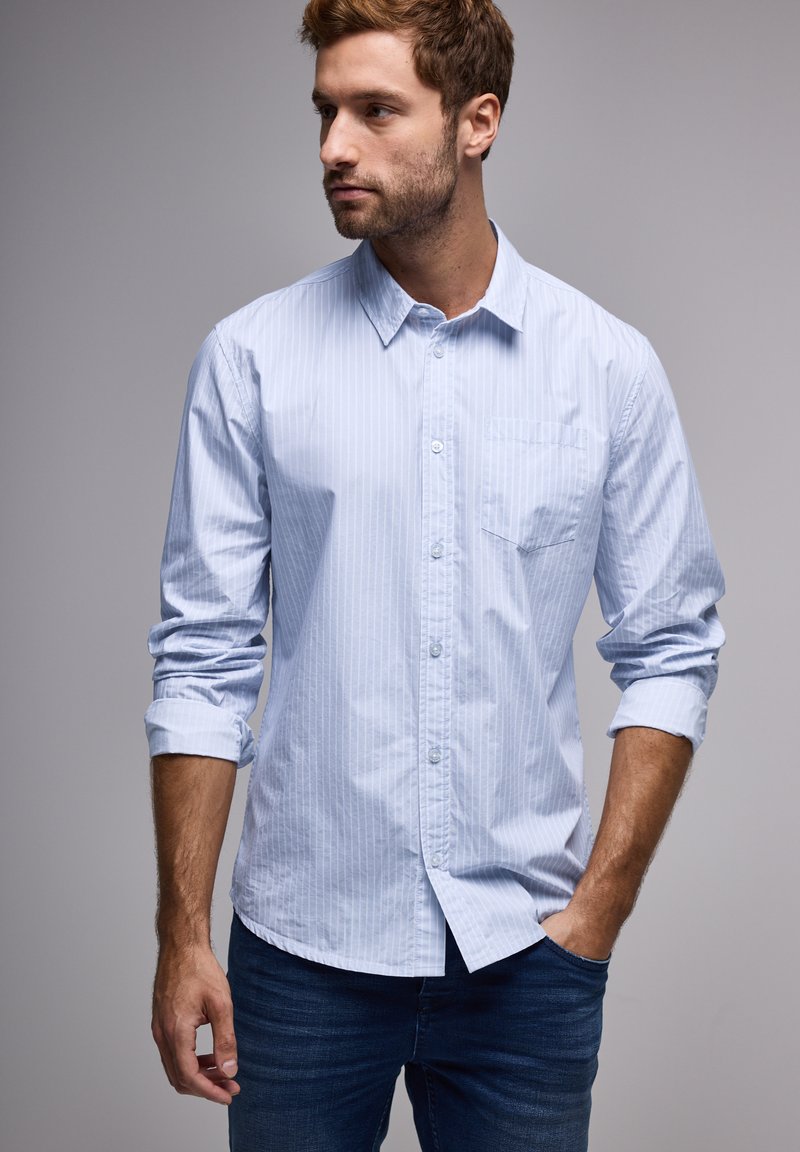 Vaaleansininen raidoitettu napillinen paita puuvillakankaasta, jossa on pitkät hihat ja rintatasku, yhdistettynä tummansinihin farkkuihin.