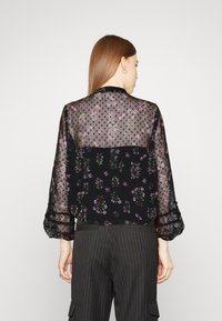 Blusa nera floreale con sovrapposizione trasparente a pois, maniche lunghe con polsini arricciati, decorata con motivi floreali rosa e verdi. Abbinata a pantaloni a righe.