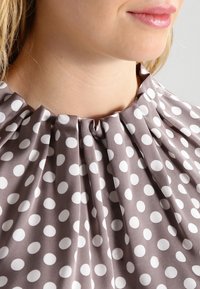 Nærbillede af en kvindes hals og hage iført en brun bluse med hvide prikker og samlede plisseringer ved kraven.