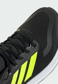 Sort atletiske sko med en mesh-overdel, lyse gule detaljer og matchende snørebånd. Letvægtsdesign med en tekstureret sål for godt greb.