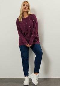 Lohikäärme-neulepusero hopeasomisteilla, yhdistettynä tummansinisiin farkkuihin ja valkoisiin lenkkareihin, seisoo neutraalia seinää vasten.