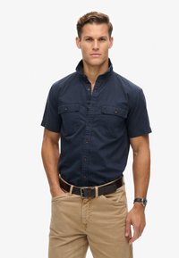 Camisa de manga corta azul marino oscuro con cuello abotonado y dos bolsillos en el pecho. Llevada con pantalones beige claros y un cinturón oscuro. Diseño sencillo.