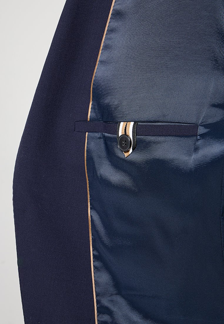 Innenseite einer Marinejacke mit glattem Satinfutter, beigem Paspel und einer geknöpften Leistentasche mit einem gestreiften Stoffabzieher.