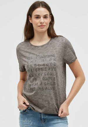 Young woman with straight brown hair wears gray short-sleeve shirt with positive phrases and blue jeans, standing against a plain background.