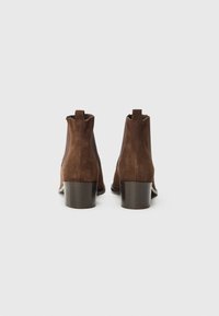 Botines de ante marrón con punta ligeramente afilada, tacón bajo en bloque y lengüetas en la parte trasera. Acentos de cuero suave en los laterales.