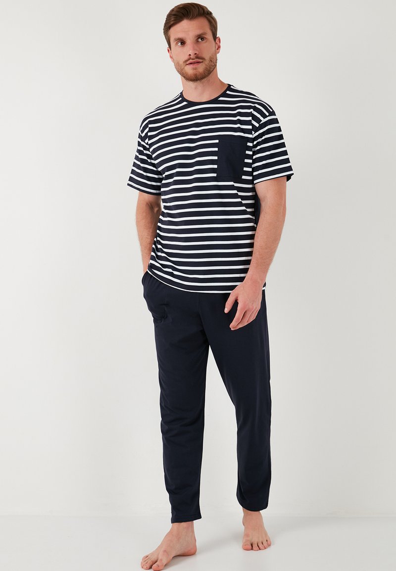 Marine gestreepte t-shirt met een borstzak, gecombineerd met uni marineblauwe pyjamabroeken. Zachte stof, comfortabele pasvorm en korte mouwen. Model op blote voeten.