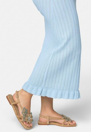 Pieds portant des sandales beiges ornées de grandes décorations florales, associées à une jupe bleu clair côtelée, longueur cheville, avec un ourlet à volants.