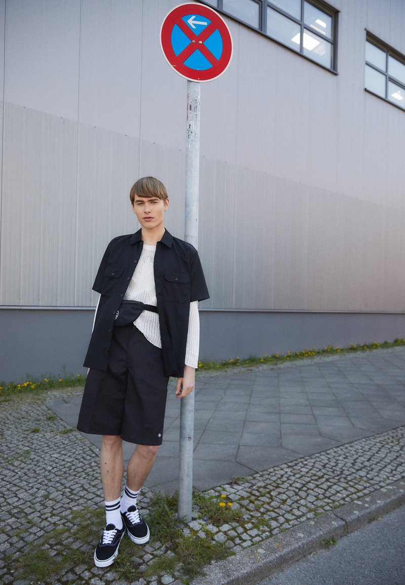 Black short-sleeve shirt with pockets over a white long-sleeve knit top, paired with black shorts and black sneakers. Signpost in background.