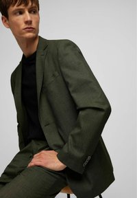 Blazer vert foncé en tissu texturé, avec une coupe slim, une poche poitrine simple et des poignets à boutons. Associé à une chemise noire.