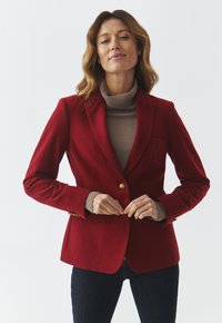Punane villane bleiser, millel on kohandatud lõige, üks kuldne nupp ja broderi detail taskul. Stylitud beezist kõrge kaelusega särk ja tumedad teksad.