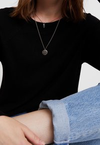 Svart långärmad topp i slät tyg; lager på lager av silverhalsband med runda och ovala hängsmycken; ljusblå jeans med uppvikta ben och mjuk textur.