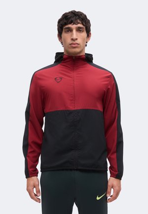 Jonge man draagt een rood-zwarte sportjas met rits en hoge kraag en donkere sportbroek met een geel logo op het linkerbeen.