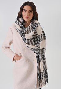 Écharpe à carreaux noir et blanc, en tissu doux, avec des bords frangés, drapée sur un manteau rose clair texturé.