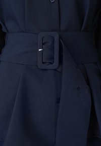 Ζώνη από σκουρόχρωμο ναυτικό ύφασμα με ορθογώνια αγκράφα καλυμμένη με ύφασμα, στερεωμένη γύρω από τη μέση του αντίστοιχου ρούχου, με ορατό πέτο κουμπιών από κάτω.