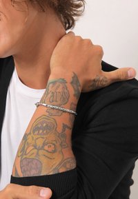 Silver bracelet featuring a braided design, worn on a tattooed forearm, contrasting with a black sweater and a white shirt underneath.