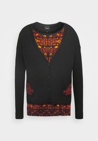 Schwarze Strickjacke mit tiefem V-Ausschnitt und Knopfleiste, mit roten Spitzenakzenten an den Taschen. Darunter ein gemustertes Top in Burgunderrot und Orange.