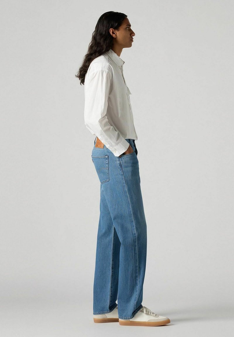 Chemise légère blanche à manches longues associée à un jean large en denim bleu et des baskets beiges, mettant en avant une coupe décontractée et une texture denim classique.