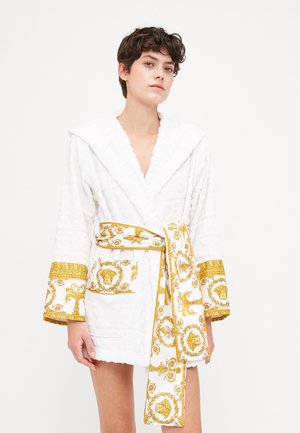Robe blanche avec un motif texturé, présentant des accents décoratifs dorés, des manches larges et une ceinture nouée ornée de motifs complexes.