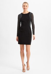 Robe noire ajustée avec de longues manches transparentes à pois, encolure ronde et silhouette slim, asociée à des talons métalliques à brides.