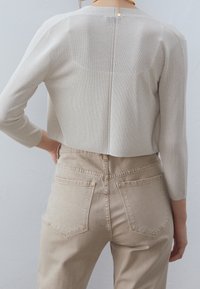 Zadní pohled na osobu v béžovém zkráceném svetru a vysokých hnědých džínech se kapsami, stojící proti jednoduché šedé zdi.