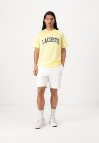 Geltonas medvilninis marškinėliai su tamsiai mėlynu "LACOSTE SPORT" spaudiniu, derinami su baltais medvilniniais šortais ir baltais batais, prieš paprastą foną.