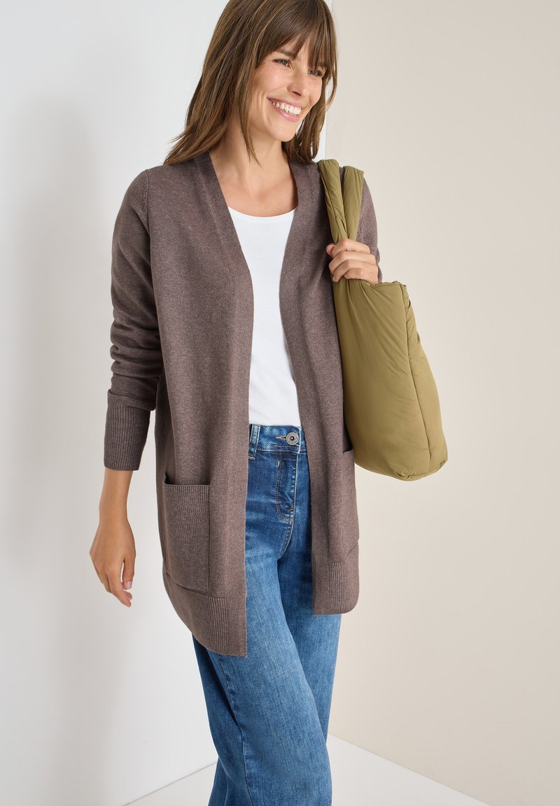 Cárdigan marrón con apertura frontal y dos bolsillos, combinado con una camisa blanca y jeans azules, sosteniendo un bolso verde oliva. Textura suave.