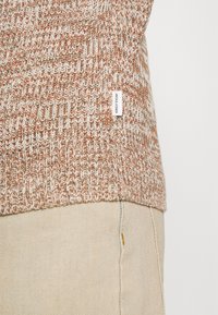 Stickad tröja med mönster i krämfärg och rostfärg. har en liten varumärkesetikett på sidan. Tillsammans med beiga byxor.