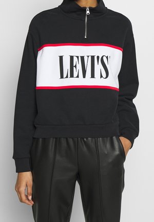 Czarno-biała bluza Levi's z czerwonym wykończeniem, kołnierzem na zamek do połowy, noszona z czarnymi skórzanymi spodniami.