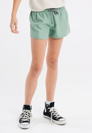 Persoon die groene sportshorts draagt met een zwart-witte koord en zwarte hoge sneakers met witte veters en zolen.