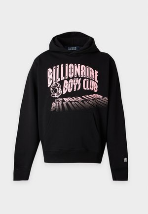 Noor mees seisab, kandes musta kapuutsiga dressipluusi roosa tekstiga „Billionaire Boys Club“, lahtisi tumedaid teksapükse ja musti tossusid ühtlasel taustal.